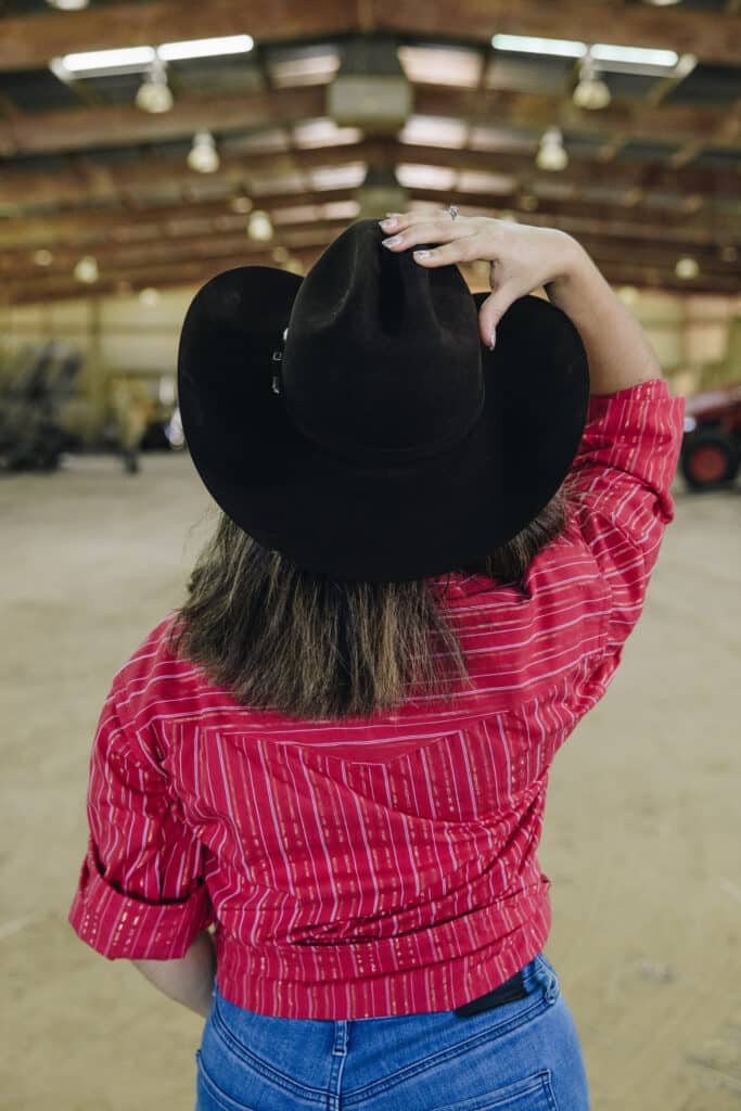 a woman wearing a red striped shirt and blue jeans holding onto a black twinstone hat in a barn