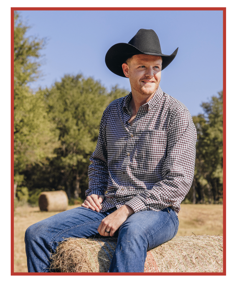 A man wearing a black cowboy hat, a checkered button-up shirt, and jeans, sitting on a hay bale in a sunny outdoor setting with trees in the background