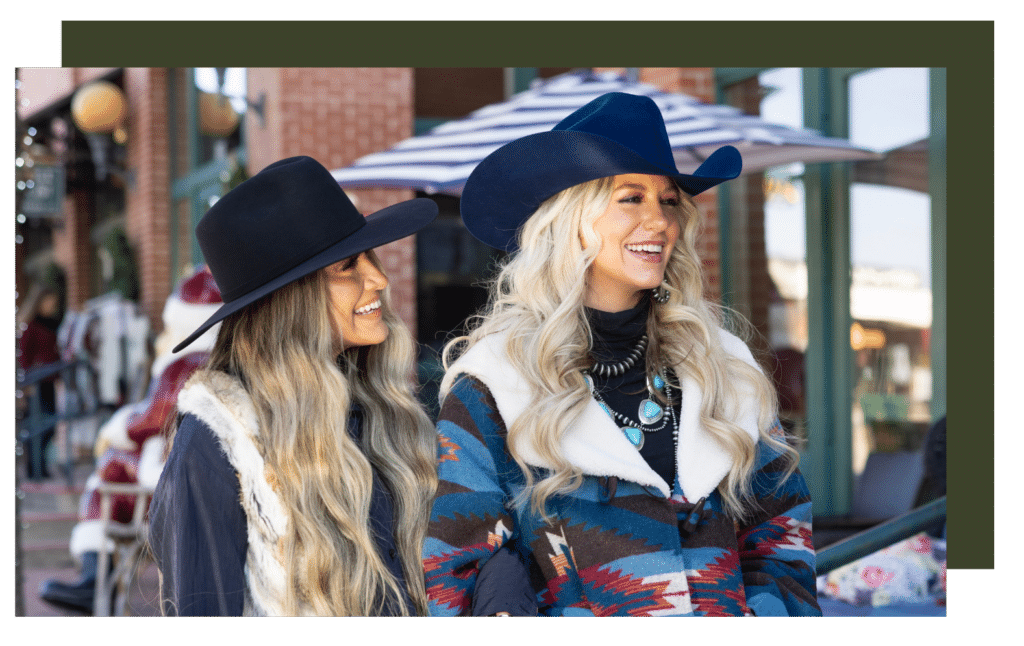 Two women wearing stylish cowboy hats, one in black and the other in blue, laughing in a festive outdoor setting.