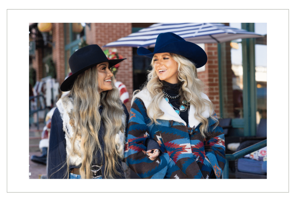 Two women in stylish cowboy hats—one in black and the other in blue—laughing in a festive outdoor shopping setting.